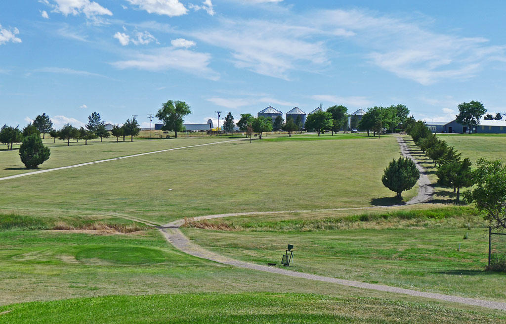 Green Golf Course lawns lined with several pine trees, Blue Sky with silver grain bins on the horizon