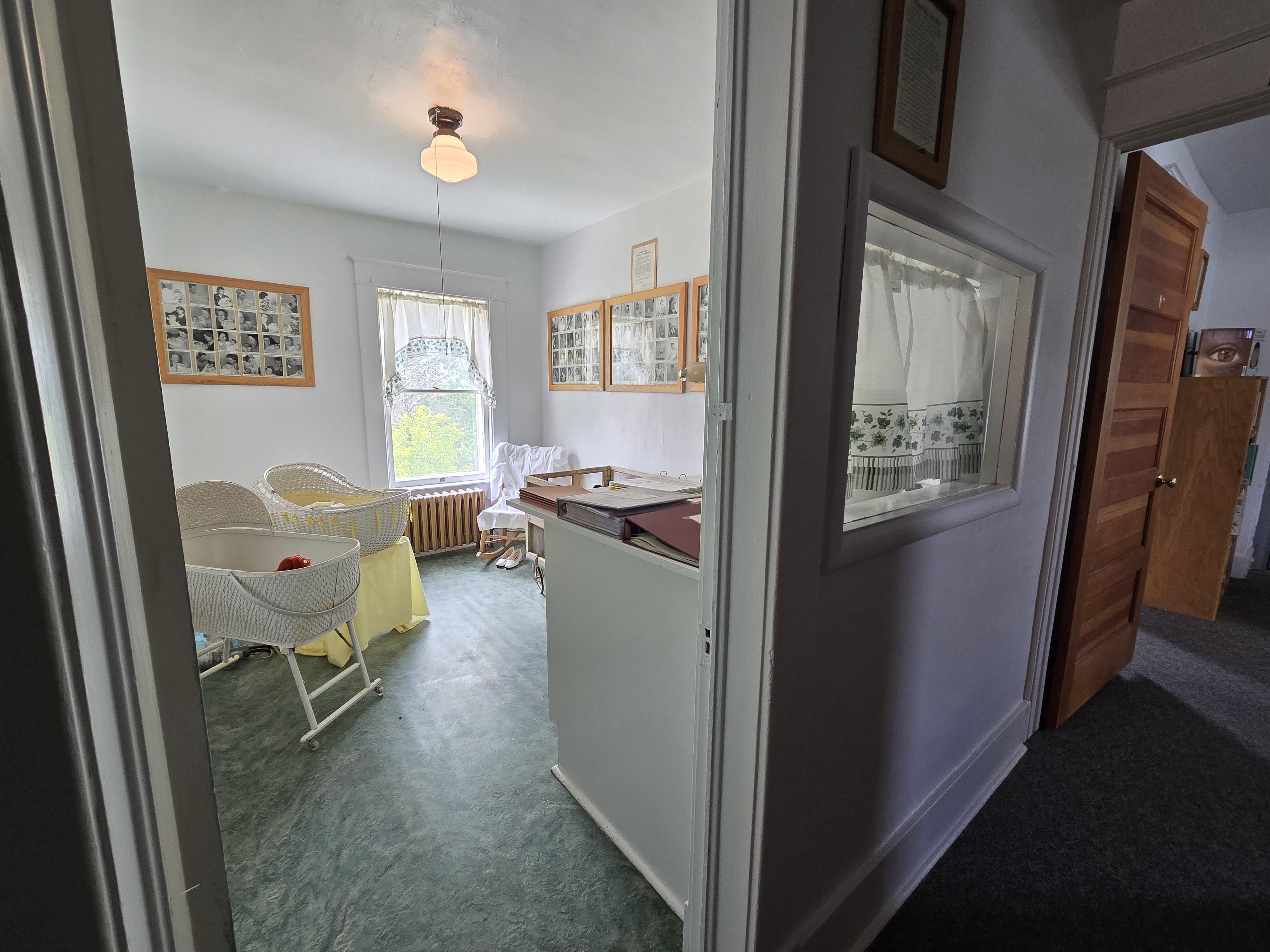 outside nursery looking in from hallway, window looking into nursery from hall way, white and green vine curtains, wooden picture frames of baby pictures, white wicker bassinettes, green linoleum flooring