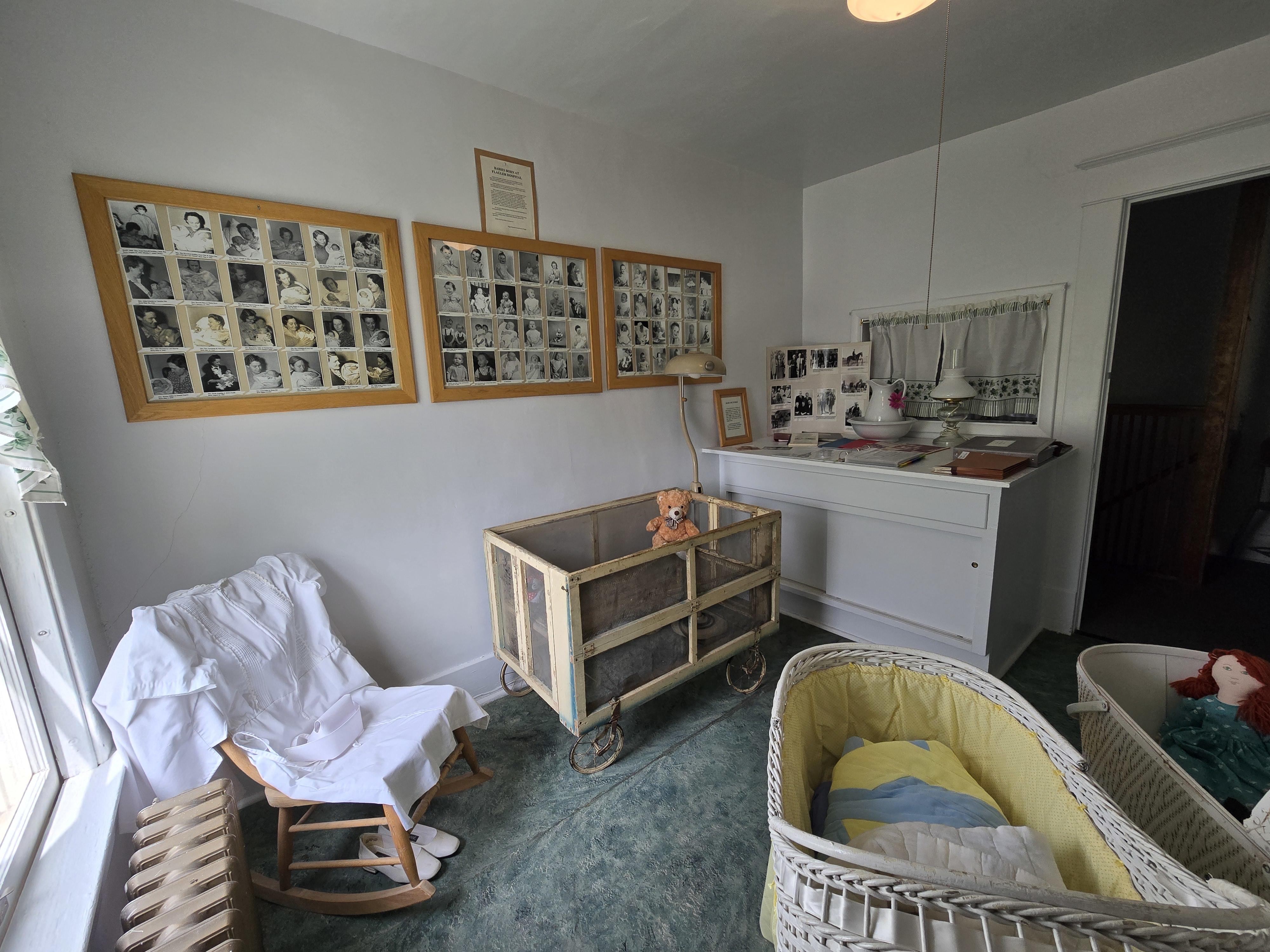 Photo of wall display of baby photos, white nurse dress and white shoes on wooden rocking chair, white and metal crib on wheels, white wicker bassinette with yellow bedding, orange teddy bear, fabric doll with brown hair and green dress